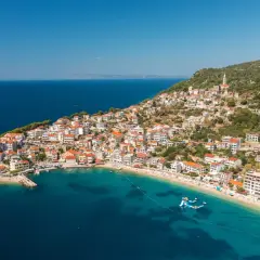 A picturesque village clings to a hillside on the Balkan coast, overlooking a turquoise bay with a sandy beach and inflatable water slides.