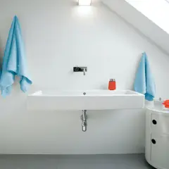 Modernt badrum med ett stilrent handfat. Två ljusblåa handdukar hänger på vänster sida, en på höger. En orange behållare och en blomma står på ett litet vitt skåp.