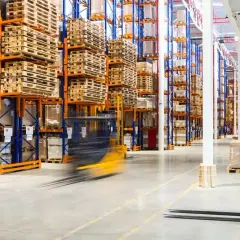 Wózek widłowy w ruchu w magazynie z wysokimi regałami pełnymi towarów i palet.