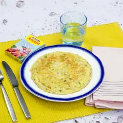 Tortilla de gambas dorada en plato blanco con borde azul, servida con cubiertos, vaso de agua y caldo de pescado.