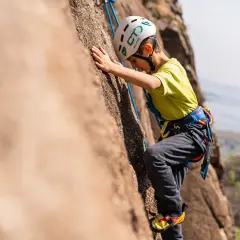 Bambino con casco e imbracatura si arrampica su una parete rocciosa. Un'avventura di arrampicata per bambini, anche se ne avesse 3 anni!