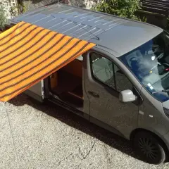Un store banne rayé orange et marron est installé sur le côté d'un fourgon gris, offrant un abri ombragé.
