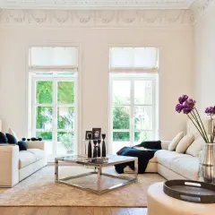 Helles Wohnzimmer im Hamptons-Stil mit beigen Sofas, einem Chrom-Couchtisch und großen Fenstern, die ins Grüne blicken.