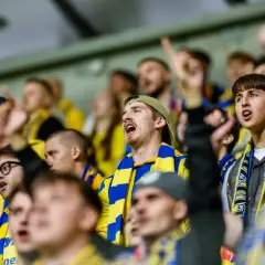 Młodzi kibice w żółto-niebieskich szalikach, wspierający warszawskie kluby piłkarskie, emocjonują się meczem.