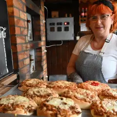 Kobieta z rudymi włosami prezentuje świeżo upieczone pizze w najstarszej pizzerii w Polsce. W tle widać ceglany piec i tablicę z logo Coca-Coli.
