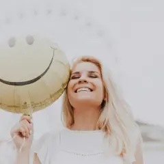 Uśmiechnięta blondynka trzymająca balon w kształcie buźki. Dobre żarty po angielsku poprawiają humor.