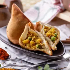 Chrupiący indyjski pierożek samosa, przekrojony na pół, ukazuje bogate nadzienie z ziemniaków i groszku. Obok łyżki z papryką i zielonymi przyprawami.