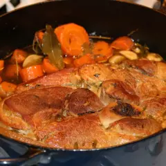 Une rouelle de porc mijote dans une cocotte avec des carottes, de l'ail et des herbes.