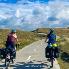 Two cyclists with panniers enjoy a scenic path through grassy dunes, perfect for Netherlands bike trips.