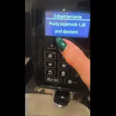 Palec z zielonym lakierem naciska przycisk na ekspresie DeLonghi. Na ekranie wyświetla się komunikat "Odkamienianie. Pusty pojemnik 1,8l pod dyszami".