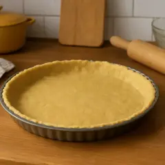 Une pâte brisée sans beurre, prête à être garnie, repose dans un moule à tarte. Un rouleau à pâtisserie et une serviette sont à proximité.