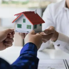 Manos intercambian una maqueta de casa, simbolizando la decisión de vender herencia.