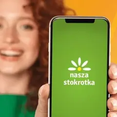 Uśmiechnięta kobieta prezentuje ekran telefonu z zieloną aplikacją "Nasza Stokrotka".