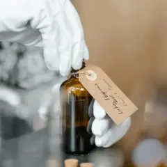Białe rękawiczki trzymają buteleczkę z ciemnym płynem. Na etykiecie widnieje napis "Linalool".