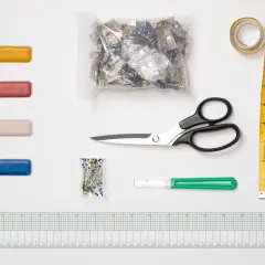A collection of essential sewing tools, including colorful clips, pins, scissors, a measuring tape, a seam ripper, and a ruler, are laid out on a white surface.