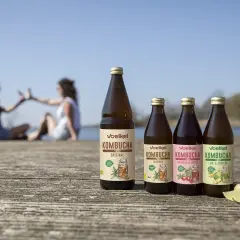 Butelki kombuchy Voelkel na drewnianym pomoście, w tle dwie kobiety wznoszące toast.