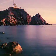 Faro en un acantilado rocoso al atardecer, con el mar tranquilo y piedras en la orilla. Parece la boca de los frailes.