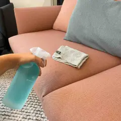 Hand sprüht Reinigungsmittel auf einen rosafarbenen Sofa-Stoff, um Wasserflecken zu entfernen. Ein Tuch liegt daneben.