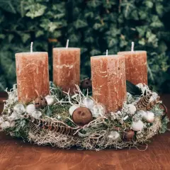 A rustic Advent wreath with four brown candles, moss, feathers, and glass ornaments on a wooden table.