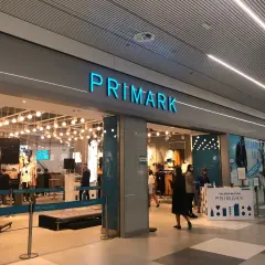 Wejście do sklepu Primark. W środku widać manekiny i ubrania. Na ladzie informacyjnej znajduje się **Primark tabela rozmiarów**.