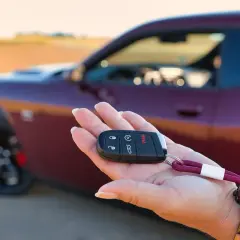Kluczyk do nowego auta w dłoni. To pierwszy krok w kupnie samochodu krok po kroku. Czerwony Dodge Challenger w tle.