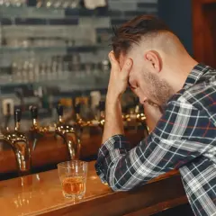Mężczyzna z rękami na głowie siedzi przy barze, obok szklanka z alkoholem. Może to być jeden z 6 objawów uzależnienia od alkoholu.