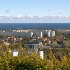 Widok na rozległe osiedla mieszkaniowe w Gdańsku, z morzem w tle. Widać charakterystyczne bloki i zieleń.