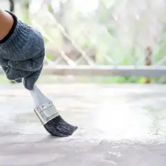 Ręka w rękawicy nakłada pędzelkiem bezbarwny preparat na betonową posadzkę. To dobry sposób, czym pomalować beton, by go zabezpieczyć.