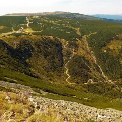Górski krajobraz z krętą ścieżką wijącą się po zboczu. W oddali widać budynki schroniska i dalsze pasma górskie. Wartość wysokości śnieżki jest tu imponująca.
