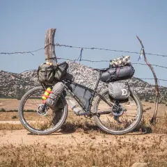 Bicicleta lista para bike packing, cargada con alforjas y bolsas, apoyada en un poste de alambre de espino, con paisaje montañoso al fondo.