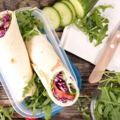 Dwie tortille z tofu i warzywami w pojemniku, obok rukola, ogórek, kiwi i maliny.