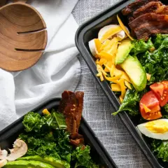 Zwei Behälter mit leckeren low carb meal prep Salaten, gefüllt mit Grünkohl, Avocado, Tomaten, Eiern, Käse und Speck.