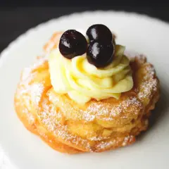 Zeppole fritte gonfie, soffice pasta con crema pasticcera e ciliegie candite, spolverate di zucchero a velo.