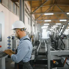 Pracownik w białym kasku przy panelu sterowania z klawiaturą. W tle hala produkcyjna z linią maszyn. Bom co to za miejsce?