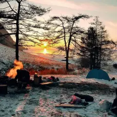Campamento invernal con fogata y tienda de campaña. La puesta de sol ilumina el paisaje nevado, un ejemplo de supervivencia y naturaleza.