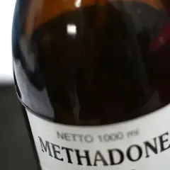 Butelka z ciemnobrązowym płynem, na etykiecie napis "Methadone" i czerwony symbol krzyża.