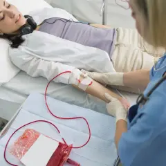 Kobieta leży w łóżku, słuchając muzyki przez słuchawki, podczas gdy pielęgniarka wykonuje przetaczanie krwi.