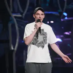 Jasiek Borkowski w czapce i białej koszulce z grafiką, trzymający mikrofon, podczas występu stand-up.