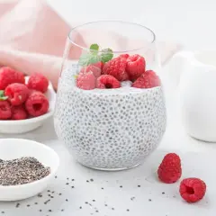 Pyszny deser z chia z malinami, udekorowany listkiem mięty. Obok miseczka z nasionami chia i świeżymi malinami.