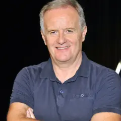 Aleksander Mikołajczak w granatowej koszulce polo, uśmiechnięty, z rękami skrzyżowanymi na piersi.