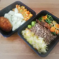 Dwa czarne pojemniki z jedzeniem: kotlet, ziemniaki, warzywa, kasza gryczana, surówka i winogrona.