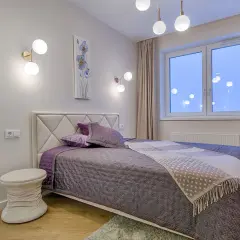 Une chambre moderne aux murs clairs, prête à peindre. Un lit douillet avec des coussins violets et une couverture à motifs. Des appliques murales design et une fenêtre lumineuse.