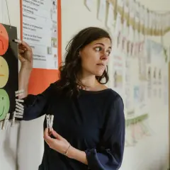 Lehrerin mit Klemmbrett und Namensschildern, die auf ein Ampelsystem mit Smiley-Gesichtern zeigen. Möglicherweise ein Hinweis auf Probleme mit der Schulleitung und Lehrern.