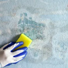 Mano con guanto blu e bianco pulisce una parete con pittura tamponato grigio e macchie di umidità.
