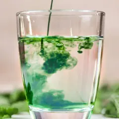 Zielony płyn rozpuszcza się w szklance wody, tworząc wiry. To naturalny chlorofil do picia, otoczony świeżymi liśćmi mięty.