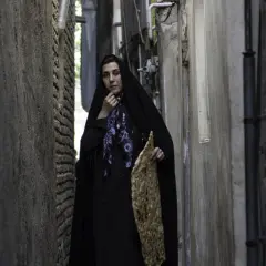 Kvinna i svart slöja med blommig scarf och ett stort bröd i handen, på en smal gränd. Kanske en scen från Lars Lennart Forsbergs film.