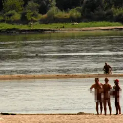 Ludzie opalają się i kąpią na plaży nudystów. Woda jest spokojna, a na brzegu widać kilka osób.