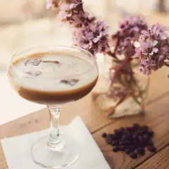 Ein cremiger Eiskaffee-Cocktail mit Eiswürfeln, daneben zarte rosa Blüten und Kaffeebohnen auf Holztisch.