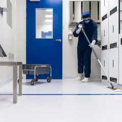 Osoba w kombinezonie i masce myje podłogę w sterylnym, clean room. W tle niebieskie drzwi i białe szafki.