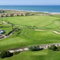 Vista aérea del campo de golf del hotel Barceló Costa Ballena, con el mar azul al fondo y edificios residenciales.